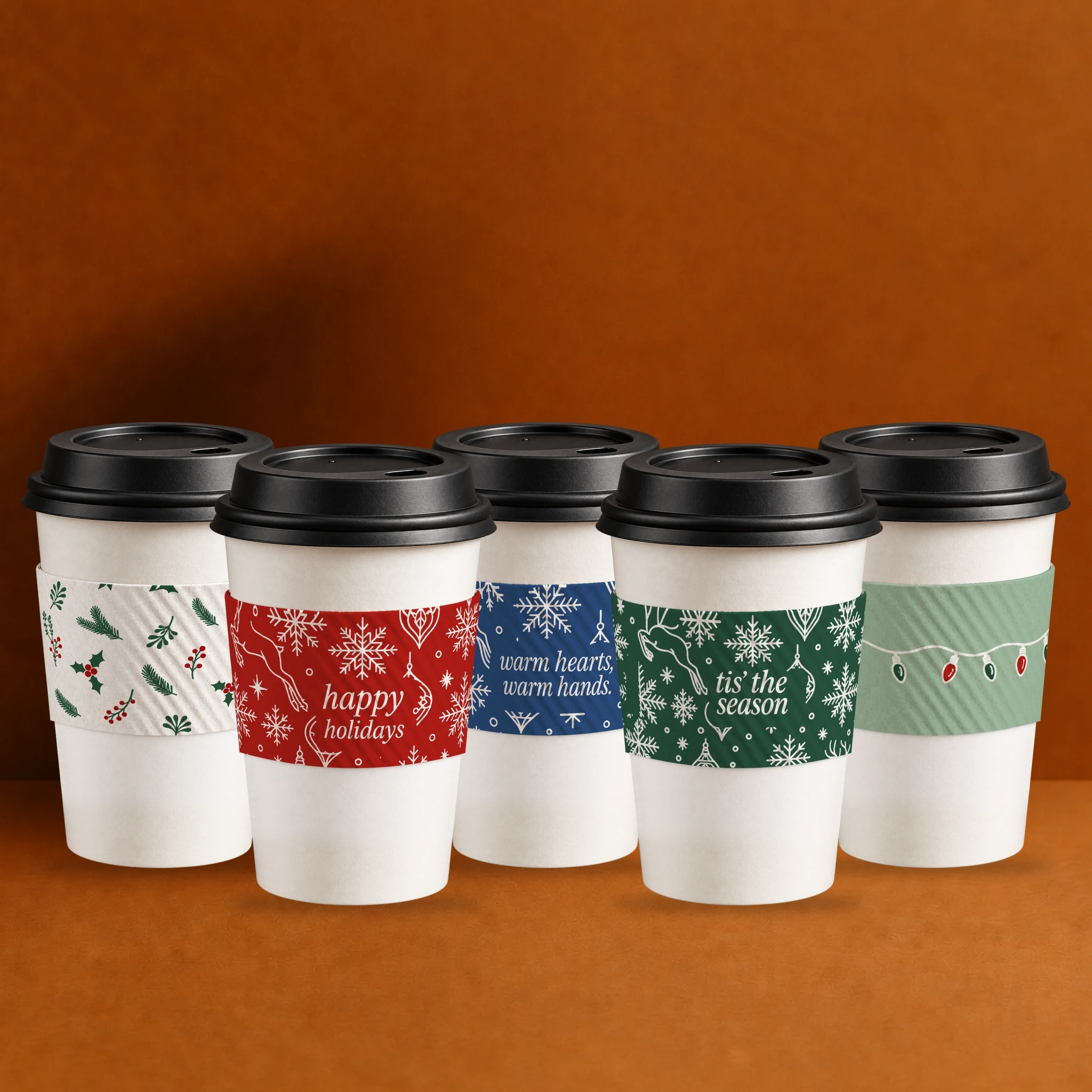 Five coffee cups wearing the Eco II Holiday 2025 Java Jacket designs: holly, happy holidays, warm hearts warm hands, tis the season and string lights